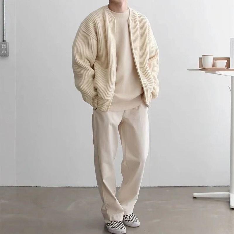 Man gekleed in een beige oversized vest, beige trui en broek, met zwart-witte geblokte sneakers, staand in een minimalistische ruimte